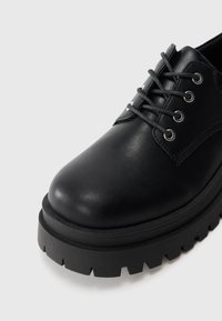 Zapato de cuero negro con punta redondeada, suela de goma negra plana con bordes dentados y ojales plateados para los cordones.