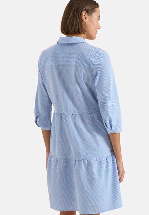 Frau trägt ein blau-weiß gestreiftes Kleid mit Dreiviertelärmeln und steht mit dem Rücken zu einem weißen Hintergrund.