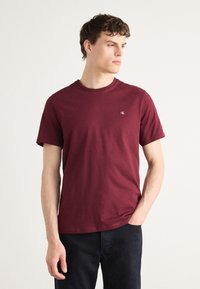 Burgunderfarvet bomulds t-shirt med rund halsudskæring og korte ærmer. Har et lille logo på venstre bryst. Båret med mørke jeans.