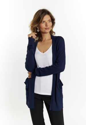 Marineblauer Strickcardigan mit langen Ärmeln, offener Vorderseite, zwei vorderen Taschen, getragen über einem weißen V-Ausschnitt-Top und schwarzen Hosen. Glatte Textur.