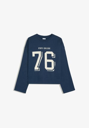 Jeune femme aux longs cheveux foncés portant un sweat-shirt "State College 76" bleu marine et un pantalon de survêtement gris clair, debout contre un mur en béton.
