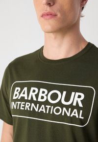 Tmavě zelené bavlněné tričko s velkým bílým logem "BARBOUR INTERNATIONAL" potisknutým v obdélníkovém rámci na přední straně.
