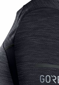 Šedý sportovní top s texturovanou tkaninou, s hladkým černým panelem a reflexním logem na spodní přední straně, které zdůrazňuje jeho inovativní design.