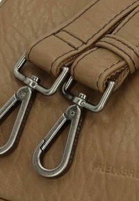 Dettaglio di una borsa in pelle tan con doppie clip metalliche e accenti di cucitura. Superficie testurizzata con logo impresso. Design complessivo compatto.