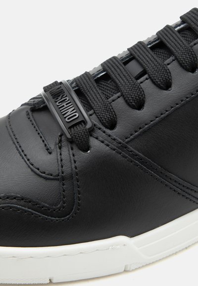 Κοντινό πλάνο μαύρου δερμάτινου αθλητικού παπουτσιού με μαύρα κορδόνια και ραμμένες λεπτομέρειες, με μια μαύρη ετικέτα κορδονιού με την επωνυμία "Moschino", ορατή λευκή σόλα.