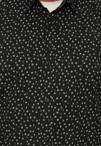 Zwarte blouse met knopen, voorzien van een patroon van witte en grijze stippen met halve maan vormen. De stof heeft een gladde textuur en is voorzien van zwarte knopen.