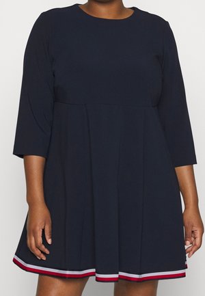 Žena v tmavě modrých šatech s tříčtvrtečními rukávy a červenobíločerveným proužkovaným lemem na spodním okraji.