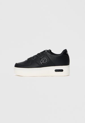 Zapatilla deportiva de cuero negro con suela blanca elevada, cordones negros y logo "RJ" en el lateral, mostrada de perfil sobre un fondo liso.