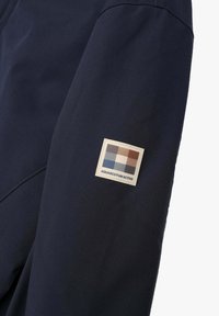 Manica di giacca impermeabile blu marina con un'etichetta di tessuto quadrata che presenta un motivo a scacchi in marrone, bianco e accenti blu.