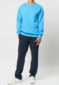 Sudadera azul clara con mangas largas, cuello redondo y un pequeño logo verde. Combinada con pantalones azul marino oscuro y zapatillas blancas.