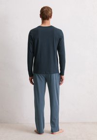 Man staat blootsvoets met zijn gezicht naar de muur, draagt een lange mouwen donkere top en een patroon met blauwe pyjamabroek op een beige vloer.