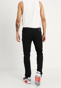 Black denim jeans with a slim fit, featuring rear pockets and a leather label. The model wears a white tank top and red-accented sneakers.