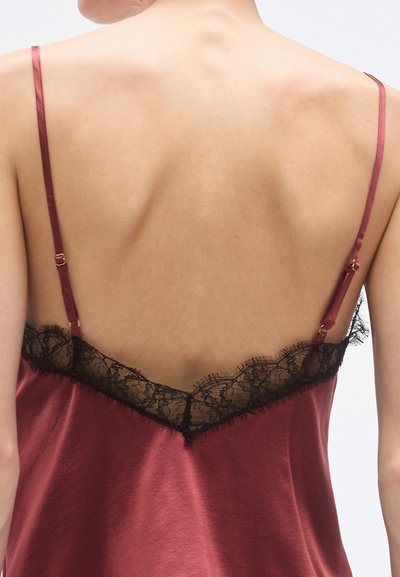 Sarkana satīna kamzole ar melnu mežģīņu apdari gar izgriezuma malu, tievām regulējamām siksniņām un gludu faktūru.