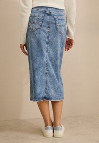 Denim-Bleistiftrock in Hellblau, knielang mit hinteren Taschen und einer Nahtdetail. Getragen mit weißen Sneakers vor einem neutralen Hintergrund.