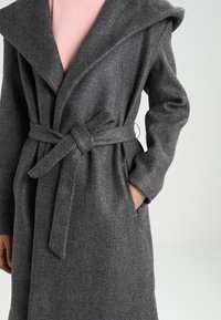 Abrigo largo de lana gris con solapas anchas, cintura ceñida y bolsillos. Presenta una textura suave y un corte relajado con un diseño minimalista.