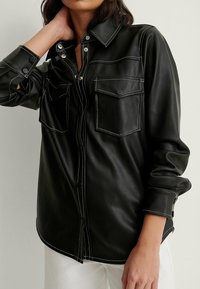 Svart läder skjorta med tryckknappar och två bröstfickor. Har kontrasterande vit sömnad och en klassisk krage, med en normal passform.