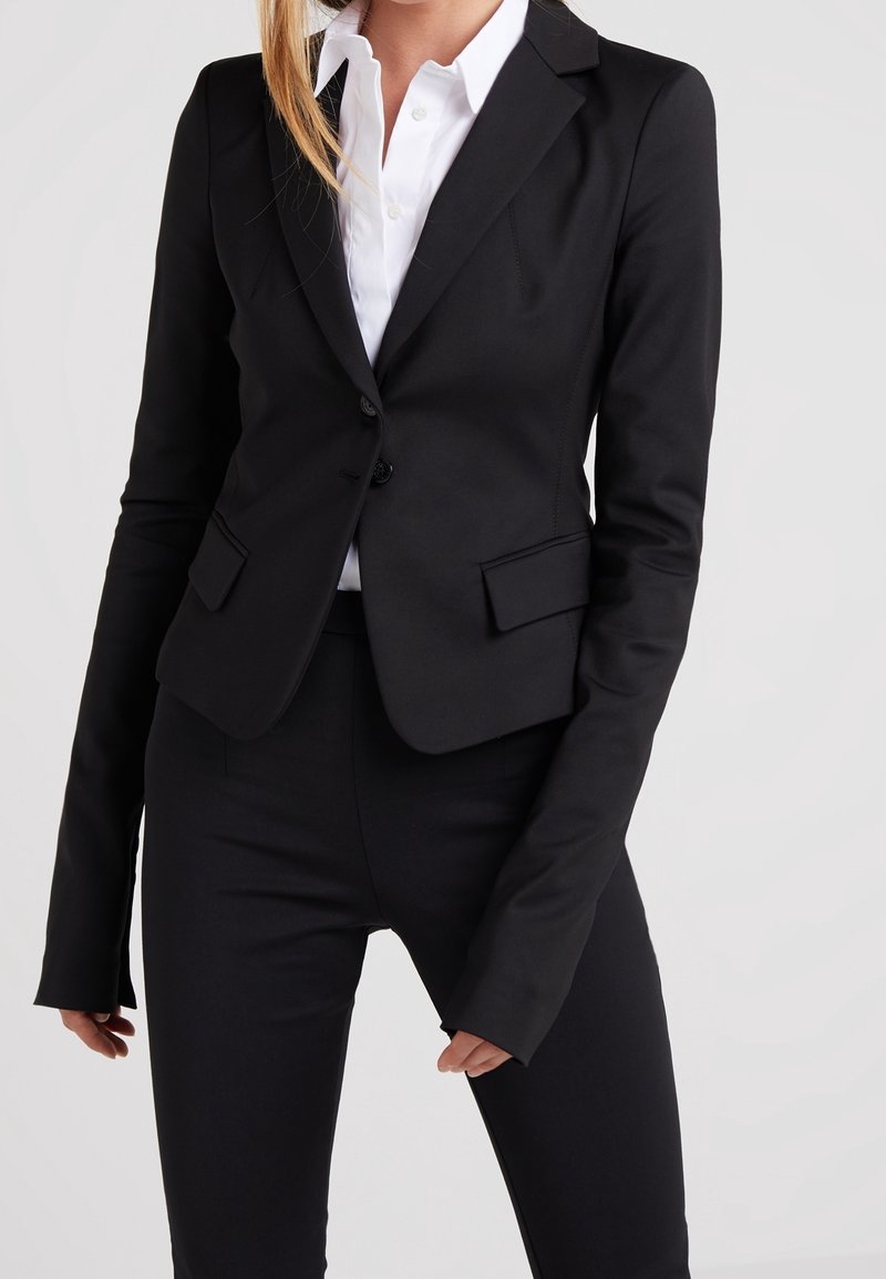Černý pološitý blazer s jedním knoflíkem, klopami a dvěma předními kapsami. V kombinaci s odpovídajícími černými kalhotami.