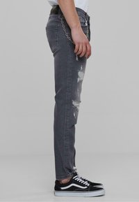 Grå denimjeans med slitte detaljer, kjedeaccessoir på livet, slim fit, og brukt med svarte sneakers med hvite detaljer.