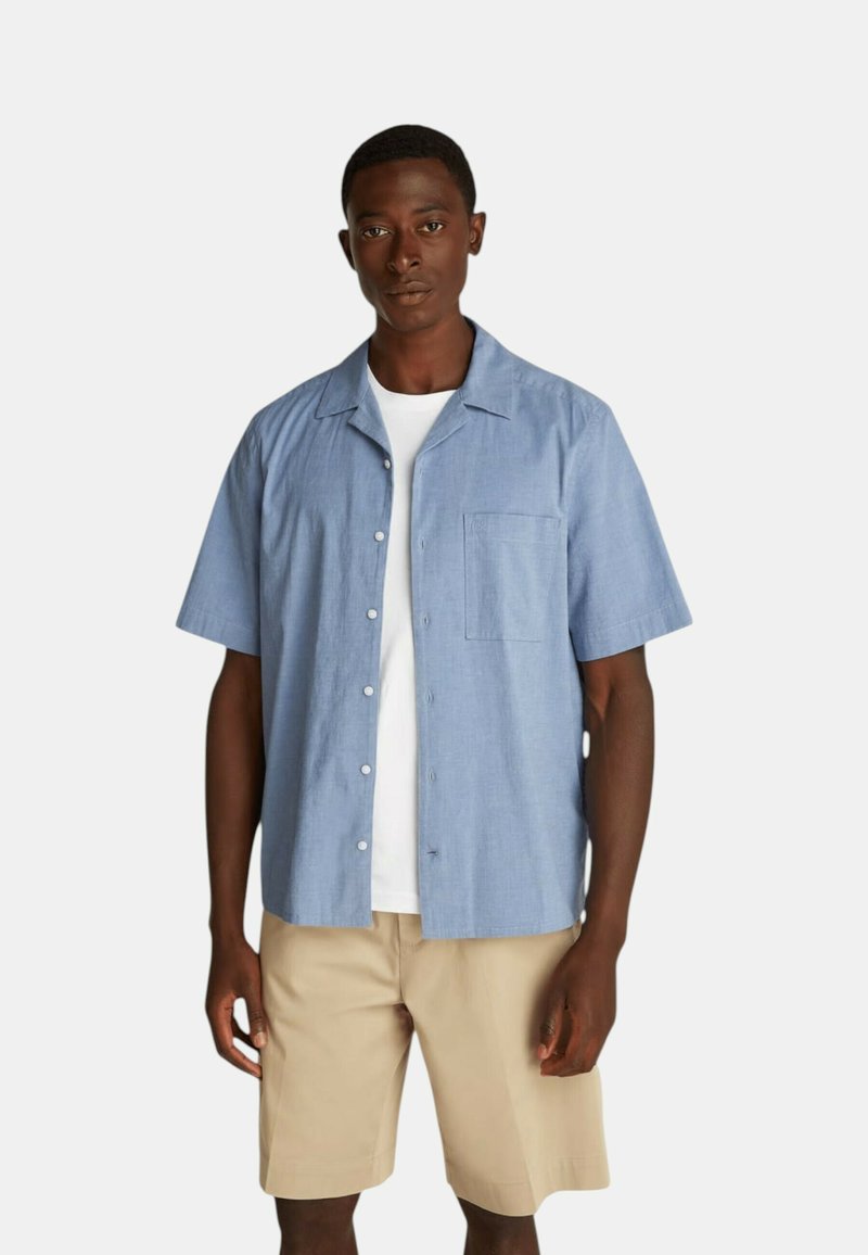 Camicia azzurra a maniche corte con bottoni in tessuto leggero, con taschino sul petto, indossata sopra una T-shirt bianca e abbinata a pantaloni corti beige.