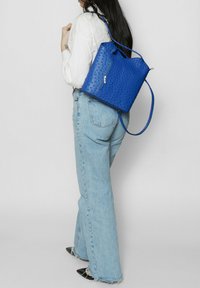 Sac à dos bleu en cuir perforé avec une fermeture éclair, des bretelles amovibles et une forme rectangulaire structurée. Porté sur le dos avec un jean bleu clair.