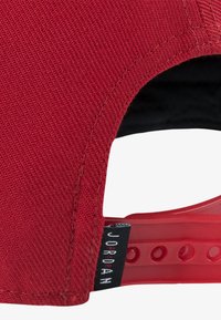 Cappellino di tessuto rosso con superficie testurizzata, visiera curva e cinturino trasparente regolabile. Presenta un'etichetta nera con la scritta "JORDAN" in bianco.