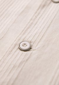 Bouton blanc cousu sur un tissu beige avec des plis cousus verticalement, partie d'un vêtement.