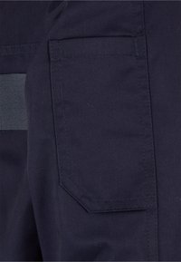 Gros plan d'un pantalon en tissu foncé montrant une poche latérale rectangulaire avec des coutures visibles et une sangle grise sur le côté gauche.