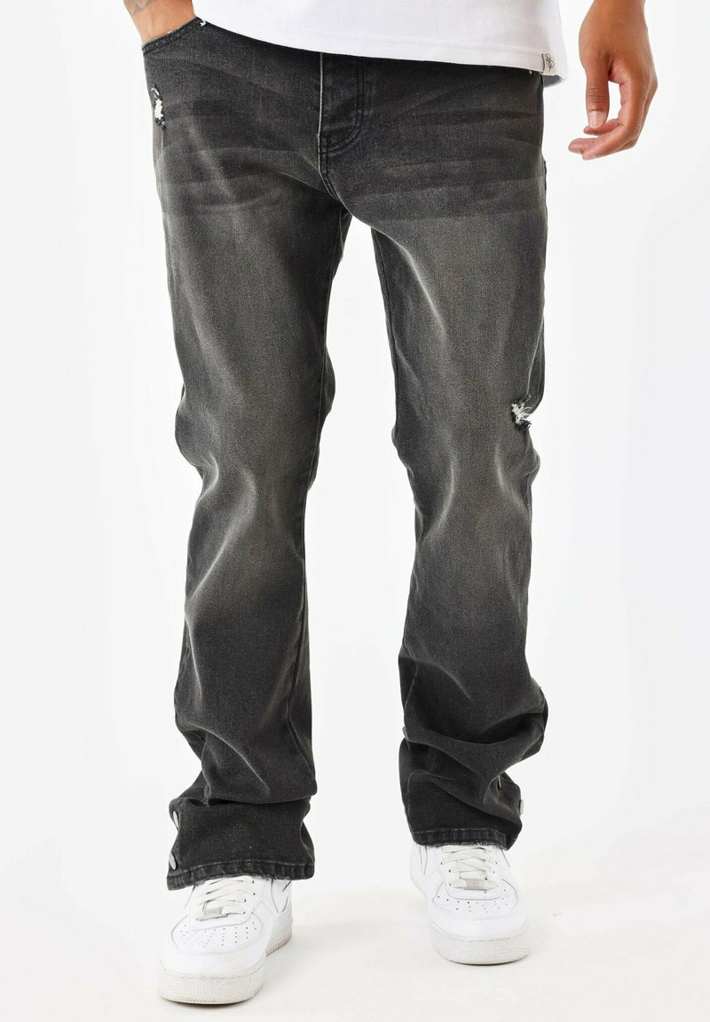 Jeans de mezclilla negra con un acabado desgastado, con un parche rasgado en el muslo, corte holgado y diseño de cinco bolsillos. Zapatillas blancas complementan el conjunto.
