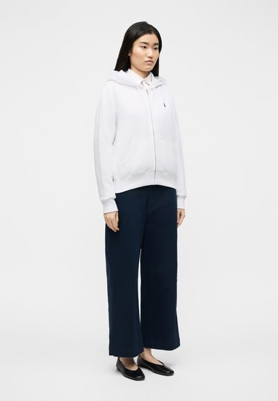 Donna in piedi che indossa una felpa bianca con zip sopra una camicia con collo, pantaloni larghi navy e ballerine nere su uno sfondo bianco semplice.