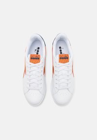 Sneaker bianchi con accenti arancioni, materiale sintetico liscio, punta tonda, profilo basso e suola testurizzata. Lacci e etichetta del marchio visibili.