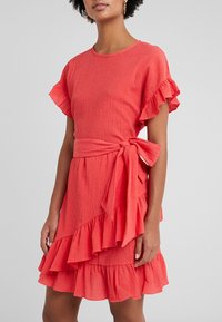 Korálové šaty s krátkými volánkovými rukávy, texturovanou látkou, zavazovacím páskem v pase a vrstvami volánků na spodním lemu.