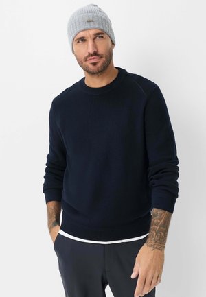 Mann trägt eine graue Strickmütze, schwarzen Pullover mit Rundhalsausschnitt und dunkle Hose, Hände in den Taschen, vor weißem Hintergrund.
