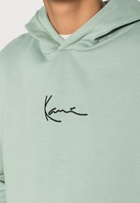 Felpa verde chiaro realizzata in tessuto morbido. Presenta una firma ricamata nera sul petto e una tasca a marsupio. Design con cappuccio e finiture a costine.