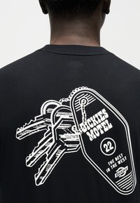 Οπίσθια όψη ατόμου που φοράει μαύρο πουκάμισο με λευκό γραφικό ενός μπρελόκ μοτέλ με την ετικέτα «Dickies Motel 22 The Best in the West.»