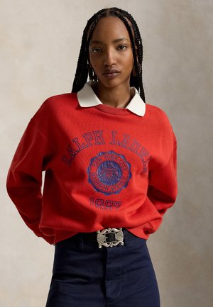 Femme aux cheveux tressés portant un sweat-shirt rouge Ralph Lauren à col blanc et un pantalon foncé avec une boucle de ceinture décorative.