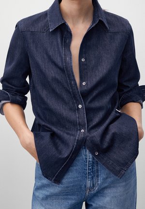 Person wearing dark denim button-up shirt with rolled sleeves and light blue jeans, hands in pockets, against neutral background.