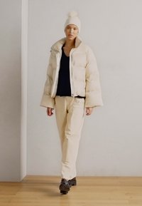 Light beige puffer jacket with a high collar, paired with a navy top and cream pants. A white knitted hat with a pom-pom. Black shoes.