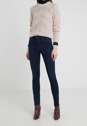 Donna che indossa un maglione rosa chiaro a trecce, jeans skinny blu scuro e stivaletti con tacco color bordeaux, in piedi davanti a uno sfondo neutro.