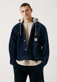 Chaqueta de mezclilla azul oscura con una capucha gris, que presenta un bolsillo en el pecho, botones a presión y un diseño clásico. Llevada sobre una camisa beige.