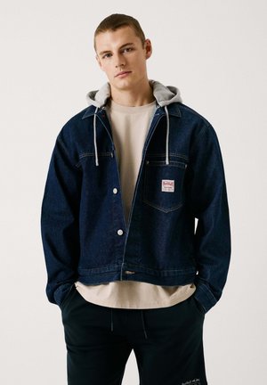 Dunkelblaue Denimjacke mit grauem Kapuzenbesatz, ausgestattet mit einer Brusttasche, Druckknöpfen und klassischem Design. Über einem beigefarbenen Hemd getragen.