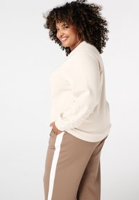 Beige sweatshirt met gestructureerde mouwen met parelachtige details, gecombineerd met bruine broek met een witte zijstreep.