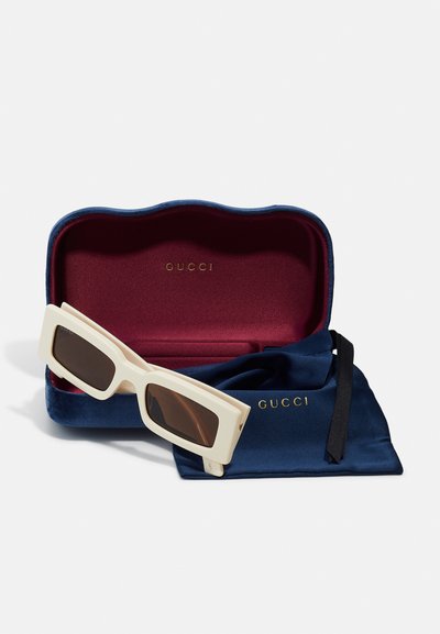Slnečné okuliare Gucci s krémovým obdĺžnikovým rámom a hnedými sklami, umiestnené v námorníckej zamatovej prípade s burgundským interiérom a logom.