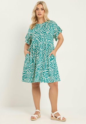 Kvinde iført en kortærmet knælængde kjole med grønne og hvide abstrakte zebrastriber, lommer og hvide sandaler med ankelstropper.
