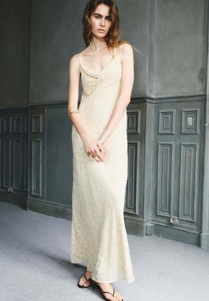 Joven mujer vestida con un vestido sin mangas de textura beige pálido y tacones negros con tiras, apoyada en una pared gris con paneles.