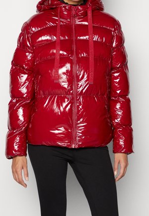 Błyszcząca czerwona puchowa kurtka z kapturem, przednim zamkiem błyskawicznym i troczkami, noszona na czarnych spodniach, widok przycięty do środka tułowia.