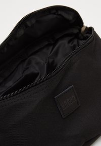 Borsetta nera in tessuto con cerniera, dotata di un interno liscio e una piccola etichetta con logo in rilievo all'esterno. Materiale resistente e design semplice.