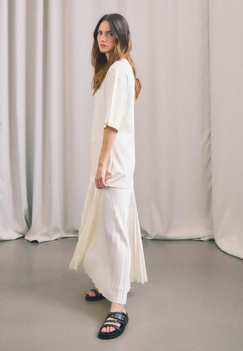 Lång, plisserad vit kjol i kombination med en kortärmad, krämfärgad topp. Svarta sandaler med cirkulära hårdvaruackenter. Neutral bakgrund.