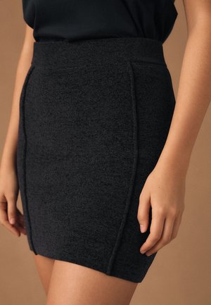 Kvinde iført en tekstureret sort strikket mini nederdel med højtaljet og lodrette sømdetaljer, kombineret med en sort top mod en neutral baggrund.