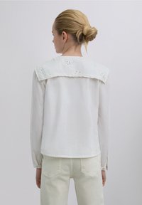 White long-sleeve blouse with an embroidered collar, featuring a smooth texture and a slightly oversized fit. Worn with light-colored pants.