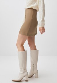 Beige Minirock mit glatter Textur, hoher Taille und seitlichem Reißverschluss. Kombiniert mit hohen, weißen Lederblockabsatzstiefeln mit hinterem Reißverschluss.